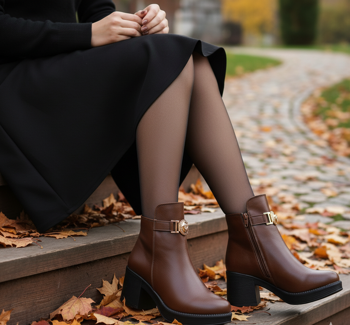Brown ankle boots with gold buckles worn by a person sitting on steps with autumn leaves.
