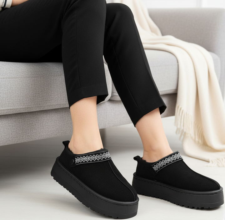 Black slip-on shoes with decorative straps worn by a person sitting on a couch.